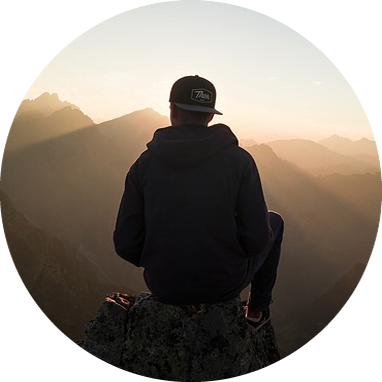 A person in a dark hoodie and cap sits on a rocky ledge, overlooking a mountain landscape at sunset with rays of sunlight streaming through the peaks.