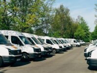 A row of white vans is parked closely together along a paved street, surrounded by green trees on a sunny day.