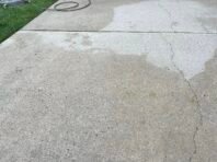 A concrete patio with a visible section that has been power washed, showing a cleaner area. In the background, there is a grill, propane tanks, a hose, and some grass and plants.